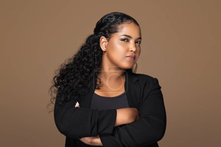 Photo for Sceptical young brazilian plus size woman standing with folded arms and looking suspiciously at camera, having some doubts, posing over brown studio background - Royalty Free Image