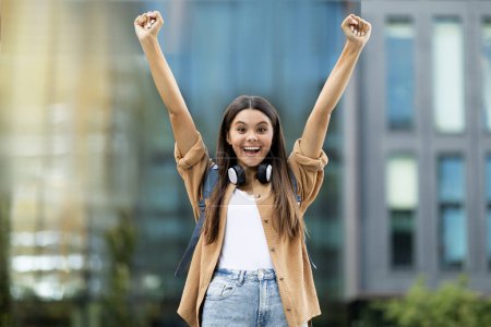 Téléchargez les photos : Heureuse jeune étudiante heureuse excitée avec sac à dos et écouteurs sans fil rasant les mains vers le haut et s'exclamant en plein air sur le bâtiment du collège universitaire, célébrant le succès dans l'étude - en image libre de droit