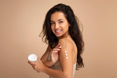 Photo for Happy attractive brunette middle eastern woman wearing beige top applying body cream on shoulder and smiling at camera, isolated on color background. Skin moisturizing, body care concept - Royalty Free Image