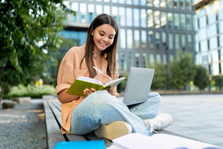 Téléchargez les photos : Joyeux jeune étudiante hispanique assise sur le banc du campus universitaire, se préparant pour la classe, utilisant un ordinateur portable et prenant des notes dans un bloc-notes. Smart lady assister à un cours éducatif en ligne - en image libre de droit