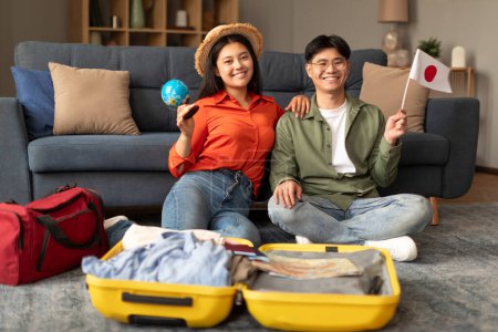 Photo for Smiling Asian Young Spouses Posing With Flag Of Japan And World Globe, Sitting Near Opened Suitcase, Planning Their Vacation Travel Or Relocation To New Country At Home Interior, Selective Focus - Royalty Free Image