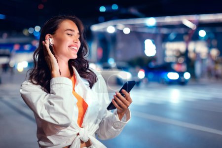 Téléchargez les photos : Jeune femme excitée marchant dans la ville la nuit et profitant de la musique dans des écouteurs avec les yeux fermés, en utilisant un smartphone et souriant, espace libre - en image libre de droit