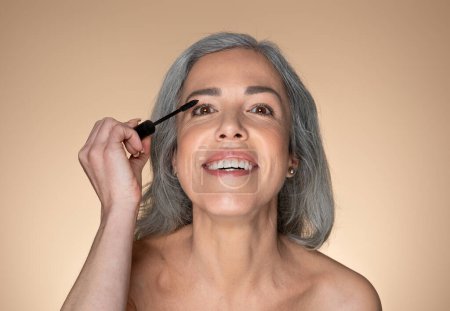 Téléchargez les photos : Portrait de femme âgée heureuse appliquant le mascara sur les cils, belle femme appréciant de faire du maquillage quotidien, posant sur fond de studio beige - en image libre de droit