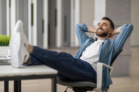 Téléchargez les photos : Pause au travail. Portrait de jeune homme relaxant au bureau avec les jambes sur la table, heureux sourire millénariste gars appuyé sur la chaise avec les mains derrière la tête, reposant sur le lieu de travail dans le bureau moderne - en image libre de droit