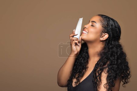 Téléchargez les photos : Joyeux brésilien femme potelée avec peau pure parfaite tenant tube de crème près du visage, posant isolé sur fond brun, vue latérale, espace de copie. Cosmétiques naturels, soins du corps et de beauté - en image libre de droit