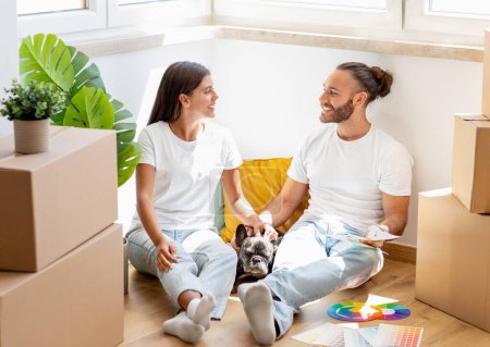 Photo for Relocation. Happy multicultural young couple sitting on floor among boxes with stuff in empty house, stroking their pet dog, have conversation, discussing interior design for new apartment - Royalty Free Image