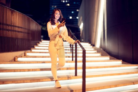 Photo for Young woman using mobile phone during walk in the night city, going down the illuminated stairs, copy space, full length. Lady using online maps or texting outdoors - Royalty Free Image