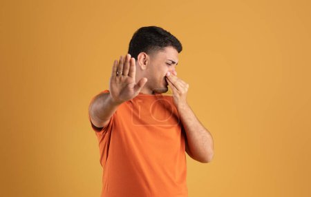 Photo for Terrible smell concept. Brazilian man closing his nose and gesturing STOP, feeling disgusted, repulsed by unpleasant odor or stinky perfume, standing over orange background - Royalty Free Image