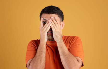 Téléchargez les photos : Homme brésilien effrayé en t-shirt orange, couvrant le visage avec les mains, debout sur fond de studio jaune, espace de copie. Problèmes, stress, émotions de peur, film effrayé, expression faciale - en image libre de droit