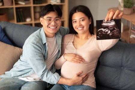 Photo for Joyful japanese couple showing sonogram photo of their baby embryo sitting on sofa indoor. Happy husband touching pregnant wifes belly while she holds ultrasound image announcing pregnancy - Royalty Free Image