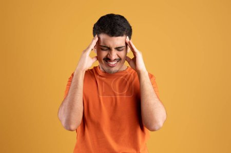 Foto de Hombre latino que sufre de dolor de cabeza severo, tener hipertensión o migraña, sentirse estresado y masajear templos sobre fondo de estudio amarillo - Imagen libre de derechos