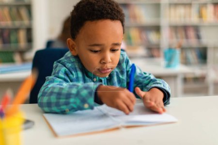 Téléchargez les photos : Étudiant afro-américain apprenant à écrire dans un copybook avec stylo, assis au bureau à l'école primaire et étudiant, espace libre. Enfants à l'intérieur de la salle de classe ou bibliothèque - en image libre de droit