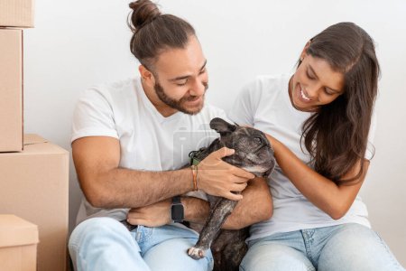 Photo for Mortgage, people and real estate concept. Happy multiethnic millennial loving couple hugging dog french bulldog, sitting on floor by carton boxes, enjoying relocation day, have break - Royalty Free Image
