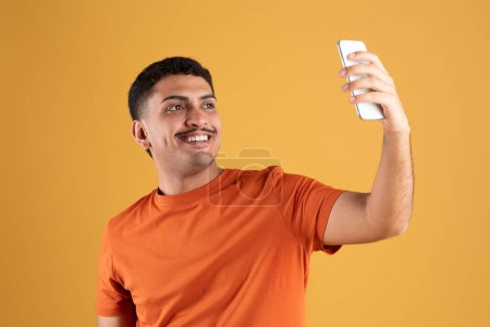 Foto de Alegre sonriente brasileño hombre sosteniendo el teléfono celular, tomando foto selfie o radiodifusión en línea, aislado en el fondo del estudio amarillo. Redes sociales, concepto de blogging - Imagen libre de derechos
