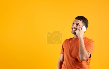 Photo for Happy brazilian man talking on cellphone, looking aside at free space, communicating having positive phone conversation, standing in studio on yellow background. Mobile communication concept - Royalty Free Image