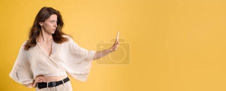 Téléchargez les photos : Confiant sérieux jeune femme caucasienne stricte dans occasionnel faire geste d'arrêt avec la main, signe de protestation, isolé sur fond de studio jaune. Lutte contre le harcèlement, le sexisme, les mauvaises nouvelles - en image libre de droit