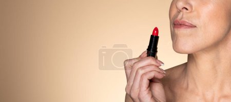 Téléchargez les photos : Femme sénior belle tenant rouge à lèvres rouge vif, posant sur fond studio beige, panorama avec espace de copie. Maquillage féminin, produits cosmétiques publicitaires - en image libre de droit