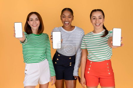 Photo for Happy pretty multiethnic young women girlfriends showing cell phones with blank screen, isolated on orange background. Cheerful ladies shopping online, recommending nice online offer, hot deal - Royalty Free Image