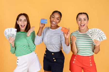 Photo for Positive cheerful three multiethnic young ladies hold money fan debit credit card, got cashback profit want buy pay on sales discounts, wear stylish outfits isolated orange color background - Royalty Free Image