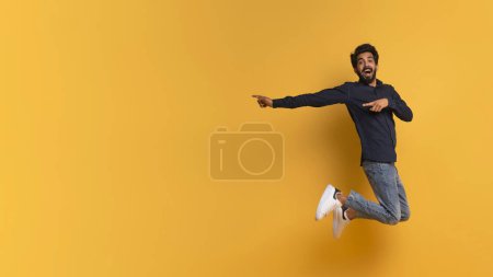 Téléchargez les photos : Super offre. Homme indien excité pointant du doigt tout en sautant dans les airs, jeune homme positif de l'Est s'amusant et démontrant de l'espace libre sur fond de studio jaune, panorama - en image libre de droit