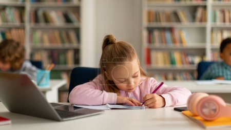 Téléchargez les photos : Écolière intelligente prenant des notes tout en e-learning sur ordinateur portable, assise au bureau et écrivant dans un copybook, camarades de classe en arrière-plan à l'école primaire - en image libre de droit