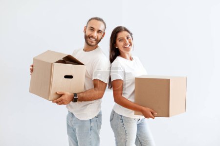 Photo for Cheerful happy millennial couple carrying cardboard boxes, packing stuff for relocation day, standing by empty white wall. Positive young man and woman moving to new house apartment - Royalty Free Image
