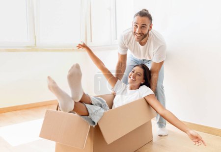 Photo for Relocation day, movement, real estate, mortgage. Happy handsome young man pushing cardboard box with girlfriend or wife cheerful woman inside, couple have fun while unpacking stuff in new house - Royalty Free Image