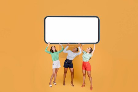 Photo for Happy excited attractive young multiethnic girlfriends carrying huge horizontal phone with white blank screen above their heads, isolated on orange background. Women showing online offer, sale - Royalty Free Image