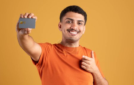 Photo for Cheerful latin man showing credit card and pointing at it, recommending bank offer, standing on yellow studio background. Check this offer concept - Royalty Free Image