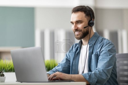 Photo for Call center operator at work. Handsome male manager wearing headset and typing on laptop while sitting at workplace in modern office, smiling employee looking at computer screen, consulting clients - Royalty Free Image