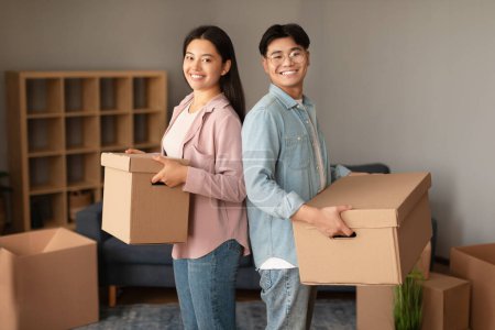 Photo for Real Estate, Apartment Rent. Cheerful Young Japanese Couple Posing In New Home, Holding Carton Moving Boxes And Smiling To Camera, Standing Back To Back At Modern House Interior - Royalty Free Image