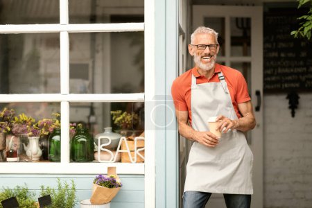 Photo for Handsome Mature Man With Coffee Standing On Terrace At His Countryside Bar, Happy Senior Small Business Owner Man Wearing Apron Posing Outdoors And Smiling At Camera, Enjoying Entrepreneurship - Royalty Free Image