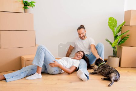 Photo for Happy young couple sitting lying on floor, have break while unpacking stuff from paper boxes, lovers enjoying their new apartment, cuddling, playing with dog. Relocation, moving concept - Royalty Free Image