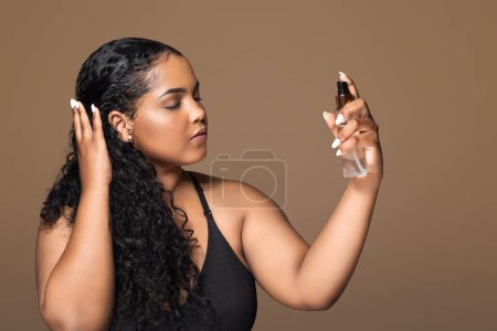 Foto de Belleza y cuidado de la piel concepto. Atractiva mujer brasileña de gran tamaño aplicando agua termal en la cara, utilizando producto cosmético, de pie sobre fondo de estudio marrón, espacio de copia - Imagen libre de derechos