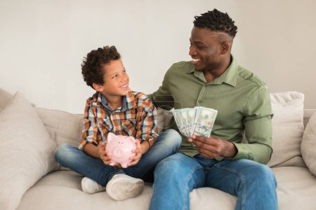 Économies personnelles. Joyeux père afro-américain et petit fils tenant le dos et l'argent en argent comptant à l'intérieur, assis sur le canapé et souriant les uns aux autres. Investissements financiers et publicité des offres bancaires