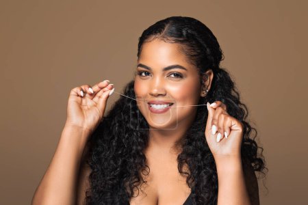 Photo for Flossing concept. Beautiful brazilian chubby woman using dental floss, smiling lady cleaning her teeth with thread while standing on brown background - Royalty Free Image