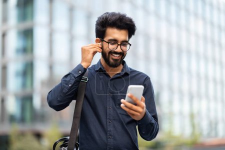 Photo for Smiling bearded young indian man wearing earbud holding phone having video call. Eastern businessman in earphone using smartphone listening music in app tech on cellphone watching videos online - Royalty Free Image