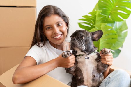 Photo for Cheerful pretty young hispanic woman sitting in carton box with puppy french bulldog dog in her hands. Happy brunette millennial lady moving to new apartment, carrying her pet - Royalty Free Image