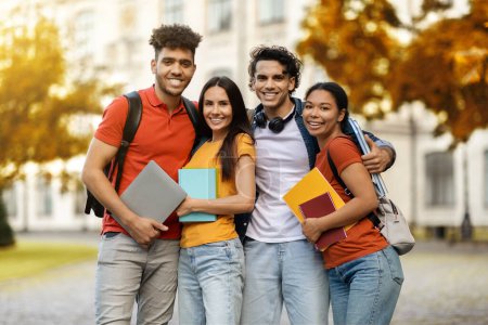 Photo for Multiethnic group of cheerful young students standing together outdoors, happy college friends embracing and smiling at camera, millennial men and women holding workbooks posing at campus, free space - Royalty Free Image