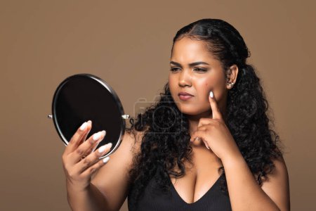 Photo for Skin disorders. Unhappy sad brazilian body positive woman looking at mirror, lady touching pimples on her cheek skin, standing over brown studio background - Royalty Free Image