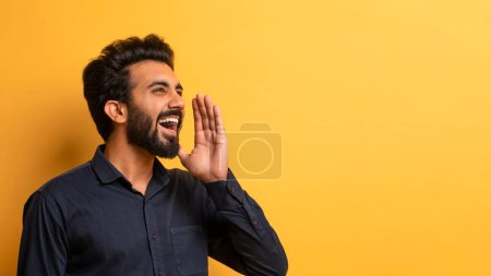 Téléchargez les photos : Jeune Indien excité avec la main près de la bouche faisant l'annonce, joyeux mâle de l'Est partageant des nouvelles ou des informations tout en se tenant debout sur fond jaune Studio, Panorama, Copier l'espace - en image libre de droit