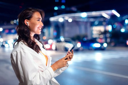 Photo for Young happy lady with earphones, standing on city street at night, listening music from smartphone while walking, illuminated city night lights on background, side view, copy space - Royalty Free Image