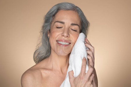Téléchargez les photos : Heureuse femme âgée caucasienne essuyant le visage avec une serviette douce et souriante, debout sur fond de studio beige. Femme faisant la routine de soins du visage le matin - en image libre de droit
