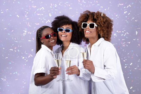 Foto de Concepto de despedida de soltera. Trío de alegres señoritas negras posando con gafas sobre fondo púrpura, brindando y celebrando juntos las fiestas, disfrutando de un vino espumoso entre los confeti que caen - Imagen libre de derechos