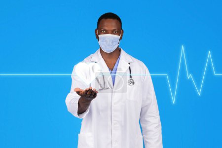Téléchargez les photos : Heureux jeune médecin afro-américain en manteau blanc, masque de protection tenir le cœur virtuel abstrait à portée de main avec cardiogramme, isolé sur fond de studio bleu. Soins de santé, médecine, traitement - en image libre de droit