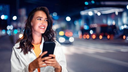Foto de Retrato de una joven excitada usando celular durante el turismo nocturno, caminando por la ciudad, señora mirando a un lado y sonriendo, panorama con espacio libre - Imagen libre de derechos