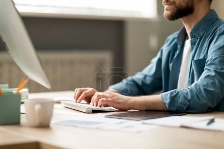 Foto de Joven barbudo sentado en el escritorio y escribiendo en el teclado de la computadora, recortado tiro de empleado masculino usando PC en la oficina, trabajando en línea, hombre de negocios contestando correos electrónicos, vista lateral, primer plano - Imagen libre de derechos