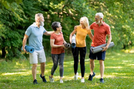 Grupo de personas mayores felices haciendo entrenamiento deportivo juntos al aire libre, hombres ancianos alegres y mujeres caminando en el parque con colchonetas de fitness en las manos, riendo y disfrutando del entrenamiento exterior, de cuerpo entero
