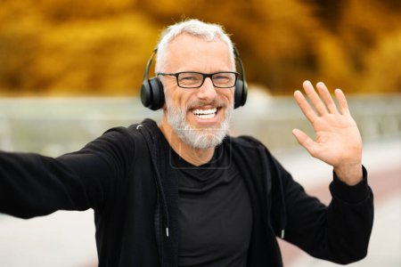 Photo for Famous fitness blogger cheerful handsome senior man wearing eyeglasses and wireless headphones taking selfie waving at camera and smiling while jogging by park at autumn, listening to music - Royalty Free Image