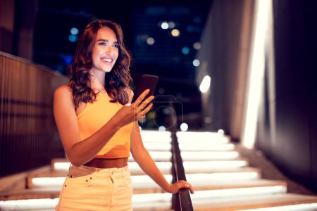 Photo for Excited young lady walking at night in the city and using cellphone, looking away and smiling while browsing online maps app using smartphone, copy space - Royalty Free Image
